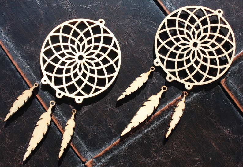 Op de afbeelding: Twee houten dromenvangers met ingewikkelde cirkelvormige ontwerpen en bevestigde veervormige hangers. De dromenvangers zijn gemaakt van lichtgekleurd hout en worden gepresenteerd op een donker, getextureerd oppervlak. De veren zijn ook van hout.