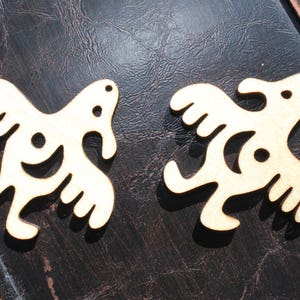 Op de afbeelding: Twee lichtbruine houten uitsparingen, elk in de vorm van een gestileerde vogel of abstracte figuur. Ze hebben gebogen lijnen en cirkelvormige uitsparingen. De achtergrond is donker en getextureerd.