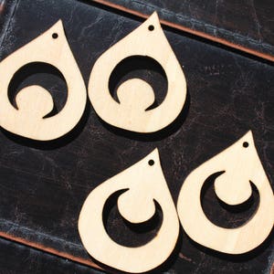 Op de afbeelding: Vier onafgewerkte houten druppelvormige oorbellen met een halve maan ontwerp. Elke oorbel heeft een klein gaatje aan de bovenkant om op te hangen. De oorbellen zijn lichtbruin en liggen op een donkerbruin, getextureerd oppervlak.