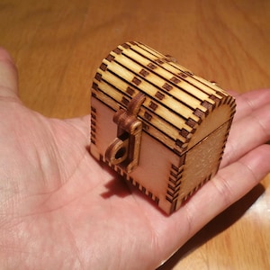 Puede incluir: Un pequeño cofre del tesoro de madera con una tapa con bisagras y un pestillo. El cofre está hecho de madera marrón claro y tiene un diseño decorativo.