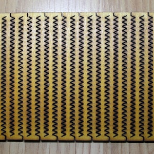 Puede incluir: Un panel de madera con un patr&oacute;n repetitivo de zigzags negros. El panel est&aacute; hecho de madera marr&oacute;n claro y tiene una superficie lisa.