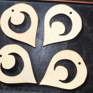 Op de afbeelding: Vier houten druppelvormige oorbellen met een uitgesneden halve maan ontwerp en een kleine stip. De oorbellen zijn lichtbruin en lijken onafgewerkt. Ze zijn gerangschikt op een donker, getextureerd oppervlak.