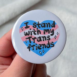May include: A white badge with the words "I stand with my Trans friends" in black script. The text is within a heart-shaped design featuring the colours of the transgender pride flag: pink, white, and blue. Small diamond shapes surround the heart.