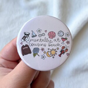 Op de afbeelding: Een ronde, lichtpaarse button met de tekst "mentally at cousins beach" in een handgeschreven lettertype. De button bevat kleine illustraties van een teddybeer, een zeilboot, een schelp, een volleybal en andere strandgerelateerde iconen.