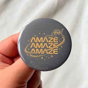 May include: A round, dark gray button with the word "AMAZE" in gold lettering, repeated three times in a circular design. A small illustration of a creature is above the word. The button is held in a hand.