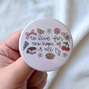 Puede incluir: Un botón redondo de color rosa claro con la frase "to live for the hope of it all" en escritura negra. El botón presenta ilustraciones coloridas de un lazo, una rodaja de limón, una concha, un sombrero, una cereza y una tarta.