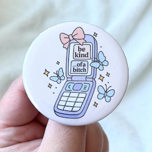 May include: A round, light purple button with a flip phone illustration. The phone displays the text "be kind...of a bitch" and is decorated with butterflies and a pink bow. The button is held in a hand.