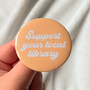 Può includere: Un bottone rotondo color pesca con la scritta "Support your local library" in corsivo bianco. Il bottone è tenuto in mano su uno sfondo bianco morbido.