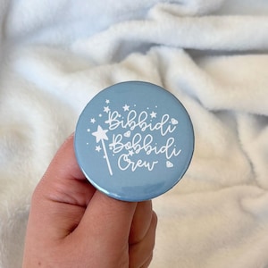 May include: A light blue round button with the text "Bibbidi Bobbidi Crew" in white script. The button features a white magic wand and star accents. The button is held against a white, fluffy background.