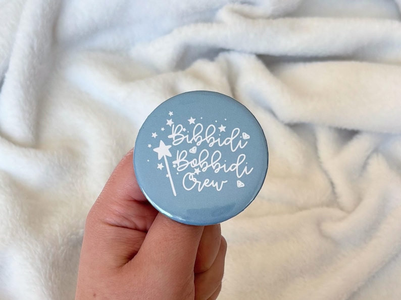 May include: A light blue round button with the text "Bibbidi Bobbidi Crew" in white script. The button features a white magic wand and star accents. The button is held against a white, fluffy background.