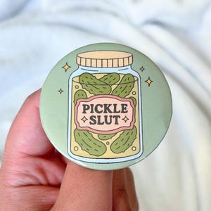 May include: A round, green button featuring a cartoon jar of pickles. The jar has a light yellow lid and contains several green pickles. The text "PICKLE SLUT" is displayed on a pink label. Four small gold stars surround the jar.