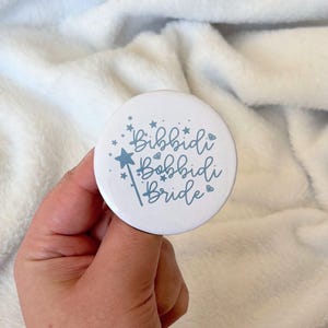 May include: A white button with the text "Bibbidi Bobbidi Bride" in a whimsical font, accented with a star and a magic wand illustration. The button is held against a soft, white background.