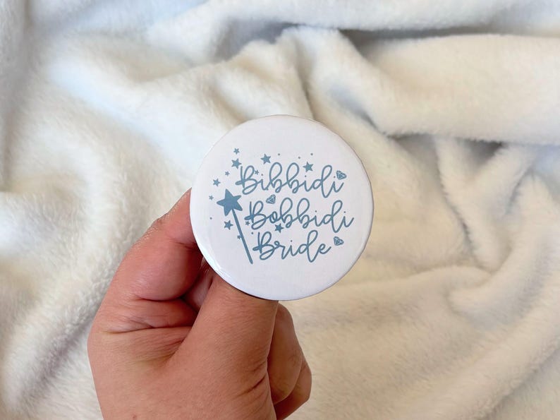 May include: A white button with the text "Bibbidi Bobbidi Bride" in a whimsical font, accented with a star and a magic wand illustration. The button is held against a soft, white background.
