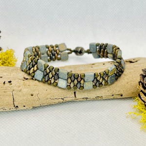 May include: A beaded bracelet featuring square and round beads in shades of blue, green, and bronze. The bracelet is displayed on a piece of driftwood, showcasing its earthy tones and intricate design. The bracelet has a magnetic clasp.