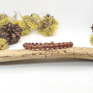 May include: A beaded bracelet with a copper-colored finish. The bracelet is made of small, rectangular beads and round beads in shades of red and copper. The bracelet is displayed on a piece of driftwood, with a white background and decorative moss and pinecone.