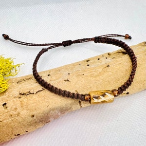 May include: A brown braided cord bracelet with a light brown wooden bead with white markings. The bracelet has a sliding knot closure.
