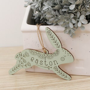 green wooden easter basket tag shaped like a bunny, personalized with the name easton. tag is decorated with intricately engraved flowers and whimsical elements. it is shown with a jute string in front of a plant.
