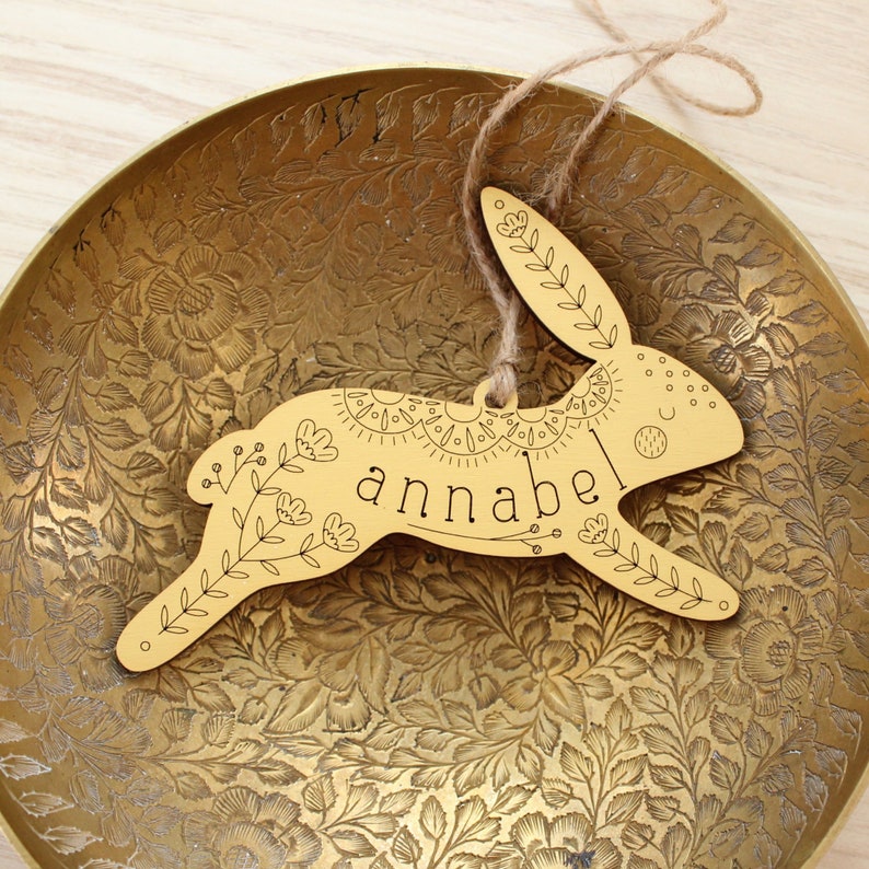 yellow wooden easter basket tag shaped like a bunny, personalized with the name annabel. tag is decorated with intricately engraved flowers and whimsical elements. it is shown with a jute string laying in a brass bowl.
