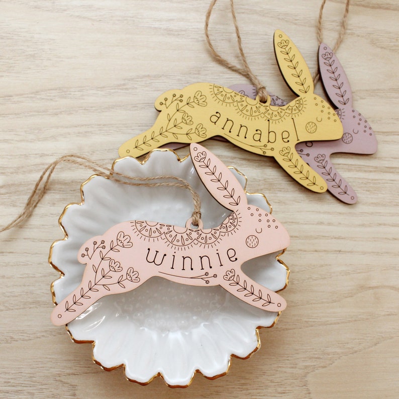 wooden easter basket tags shaped like bunnies. the one in the front is pink with the name winnie and is decorated with intricately engraved flowers and whimsical elements. in the background is a yellow bunny with the name annabel and a purple bunny.