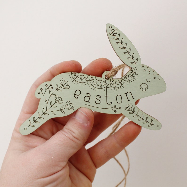 green wooden easter basket tag shaped like a bunny, personalized with the name easton. tag is decorated with intricately engraved flowers and whimsical elements. it is shown with a jute string being held in hand.