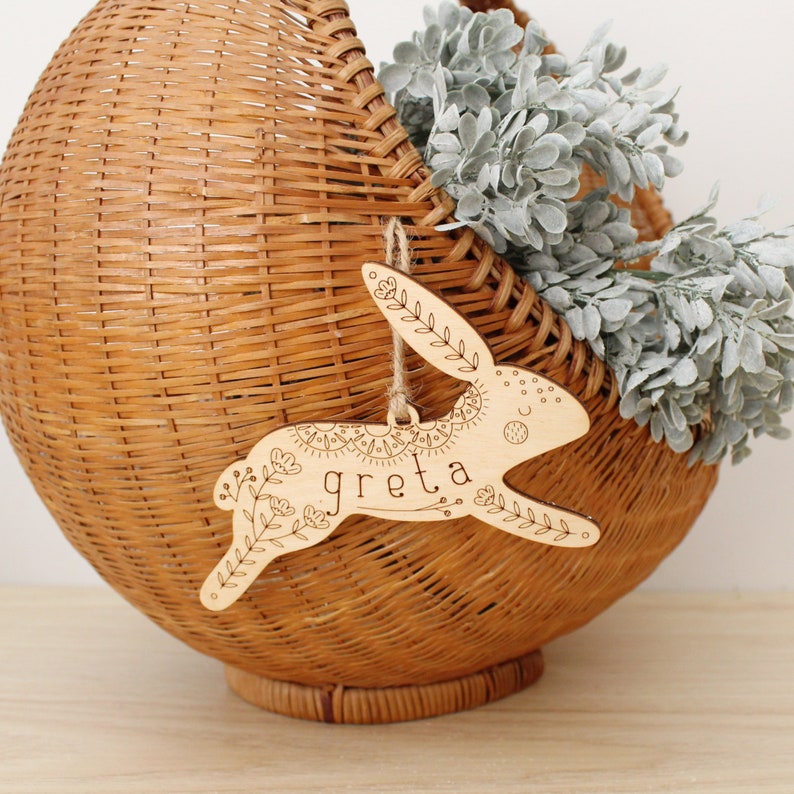 natural, light toned wooden easter basket tag shaped like a bunny, personalized with the name greta. tag is decorated with intricately engraved flowers and whimsical elements. it is shown hanging from a jute string on a basket filled with greenery.