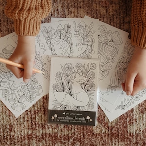 May include: A pack of black and white woodland-themed postcards featuring a fox illustration, with a black band reading "woodland friends" and "14 postcards to colour and send". Other postcards with similar illustrations are also visible.