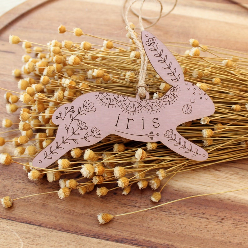 purple wooden easter basket tag shaped like a bunny, personalized with the name iris. tag is decorated with intricately engraved flowers and whimsical elements. it is shown with a jute string laying on dried flowers on a wooden plate.