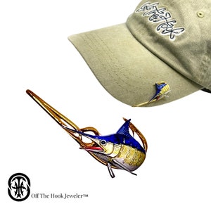 May include: A detailed, colorful marlin pin with blue, yellow, and white accents. The pin is displayed next to a green baseball cap with the text "Off The Hook" embroidered on it. The pin is a product of Off The Hook Jeweler.