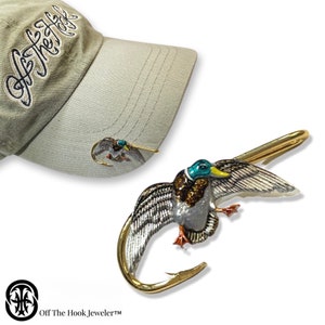 May include: A gold-colored duck with green and blue accents is attached to a fishing hook. The duck is flying with its wings spread. The hook is attached to a hat with the text "Off The Hook, Jeweler™" on the front.