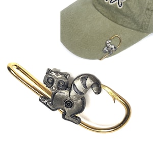May include: A gold-colored fishing hook with a raccoon design. The raccoon is holding the hook in its mouth. The hook is attached to a green baseball cap with the text "Cast the Hook" embroidered on it.