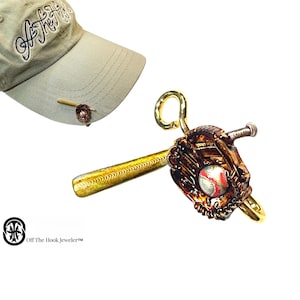 May include: A gold baseball themed hat pin with a baseball glove, bat, and ball. The pin is attached to a baseball cap with the text "Off The Hook Jewelers" on the front.