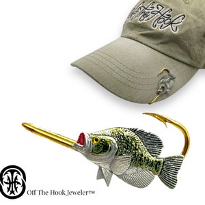 May include: A gold and silver crappie fish pin with a red mouth and green and white scales. The pin is attached to a gold hook and is worn on a beige baseball cap with the text "Off The Hook Jeweler" embroidered on the front.