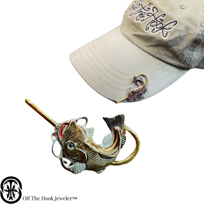 May include: A gold-tone fishing lure shaped like a catfish with a hook. The lure is attached to a brown and beige baseball cap with the text "Off The Hook, Jeweler" on the front.