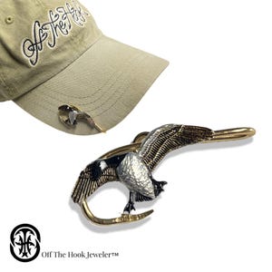 May include: A gold-tone metal hat clip featuring a detailed, black and white painted goose design. The goose is depicted in flight with its wings spread. The clip is shaped like a fishing hook. The text "Off The Hook, Jeweler™" is visible in the bottom right corner of the image.