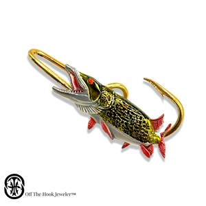 May include: A gold-colored fish hook with a detailed, realistic depiction of a pike fish. The fish is painted with green, black, and red accents. The hook is open and ready to be used.