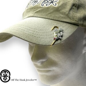 May include: A beige baseball cap with a fish hook and a small fish charm attached to the front. The fish is silver and green with a red mouth. The text "Off The Hook, Jeweler&trade;" is printed on the bottom of the image.