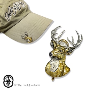 May include: A gold and silver deer head charm with a detailed design. The charm is attached to a beige baseball cap with the text "Off The Hook, Jeweler™" on the front.