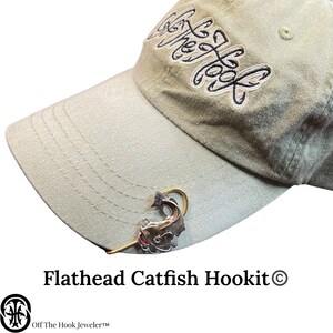May include: A silver flathead catfish hookit charm with a red lure attached to a gold hook. The charm is attached to a beige baseball cap with the text "Off The Hook Jeweler" and "Flathead Catfish Hookit &copy;" printed on the cap.