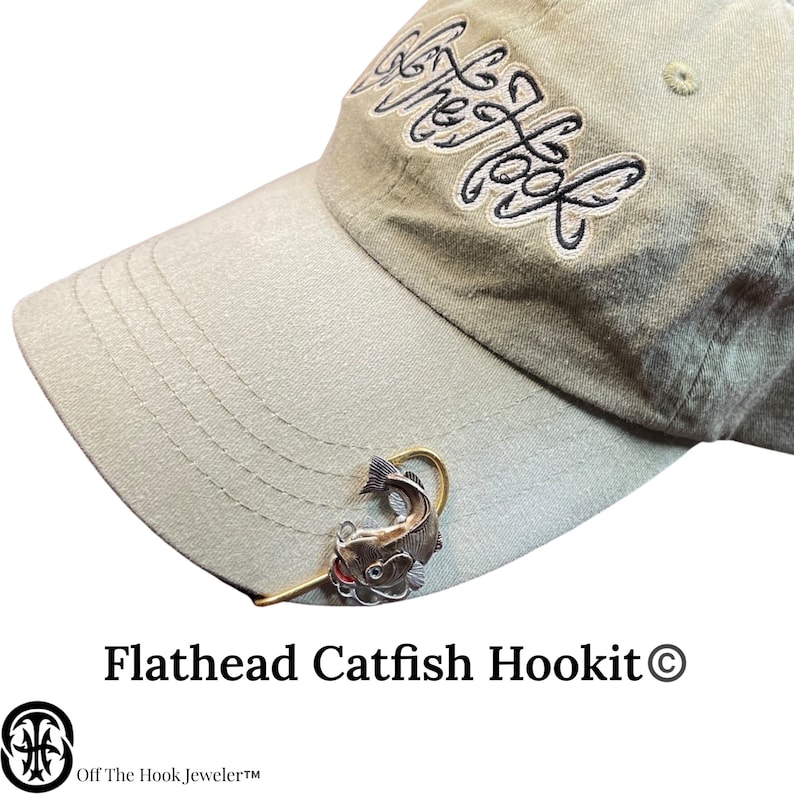 May include: A silver flathead catfish hookit charm with a red lure attached to a gold hook. The charm is attached to a beige baseball cap with the text "Off The Hook Jeweler" and "Flathead Catfish Hookit &copy;" printed on the cap.