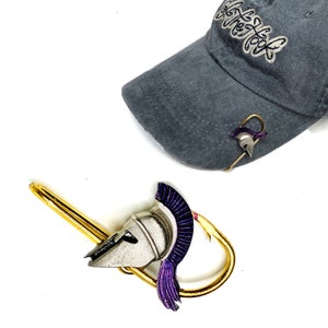 May include: A gold-colored hat clip with a silver and purple helmet design. The helmet has a purple plume. The clip is attached to a baseball cap with the text "Get the Look" embroidered on the front.