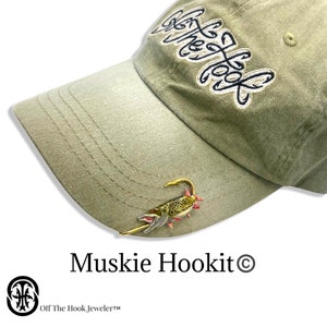 May include: A green baseball cap with the text "Off The Hook" embroidered on the front. A gold-colored fish lure is attached to the brim of the cap. The text "Muskie Hookit&copy;" is printed below the cap.