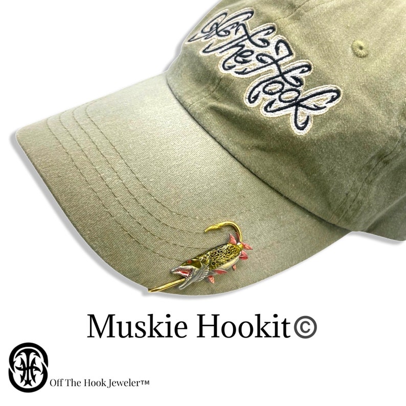 May include: A green baseball cap with the text "Off The Hook" embroidered on the front. A gold-colored fish lure is attached to the brim of the cap. The text "Muskie Hookit&copy;" is printed below the cap.