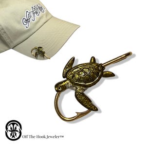 May include: A gold-colored metal sea turtle-shaped fishing hook. The hook is attached to a gold-colored metal bar. A tan baseball cap with the text "Off The Hook" is in the background. A small sea turtle pin is attached to the cap.