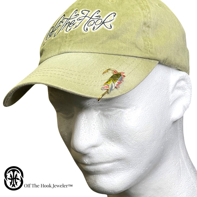 May include: A khaki baseball cap with the text "Off The Hook, Jeweler&trade;" on the side. The cap has a fish hook with a lure attached to the front.