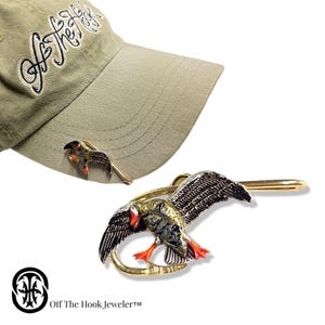 May include: A khaki baseball cap with the text "Off The Hook" embroidered in black script. A gold-toned duck pin with black, brown, and red enamel detailing is attached to the cap. A second duck pin is also visible.