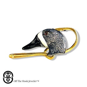 May include: A gold safety pin with a detailed enamel duck head charm. The duck has a black beak, white stripe, and gray feathers. The pin is a unique and whimsical accessory.
