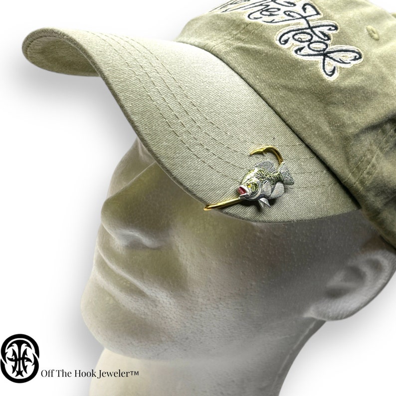 May include: A green and white fish with a gold hook is attached to a green baseball cap. The cap has the text "Off The Hook" embroidered on it.