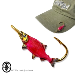 May include: A pink salmon fish pin with gold accents and a hook. The pin is attached to a green baseball cap with the text "Off The Hook, JewelerTM" on the brim.