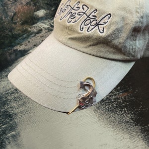 May include: A beige baseball cap with the text "Get The Hook" embroidered on the front. A gold fishing hook with a silver fish charm is attached to the brim of the cap.