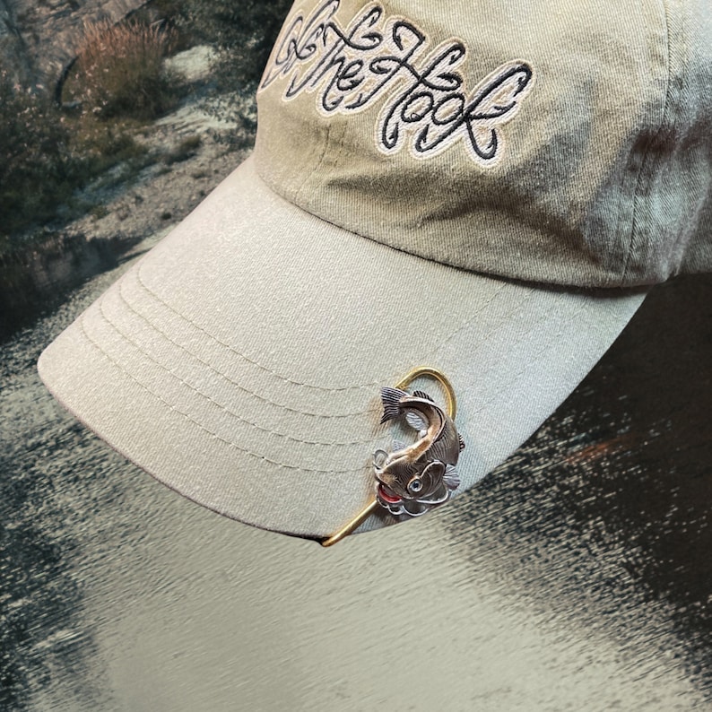 May include: A beige baseball cap with the text "Get The Hook" embroidered on the front. A gold fishing hook with a silver fish charm is attached to the brim of the cap.